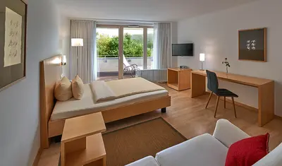Doppelzimmer mit Doppelbett, Couch, Schreibtisch und Stuhl. Fernseher und breiter Fensterfront mit Balkon