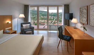 Doppelbett, Schreibtisch und Stuhl sowie Ledersessel in einem hellen Doppelzimmer. Breite Fensterfront mit Balkontür und Blick ins Grüne.