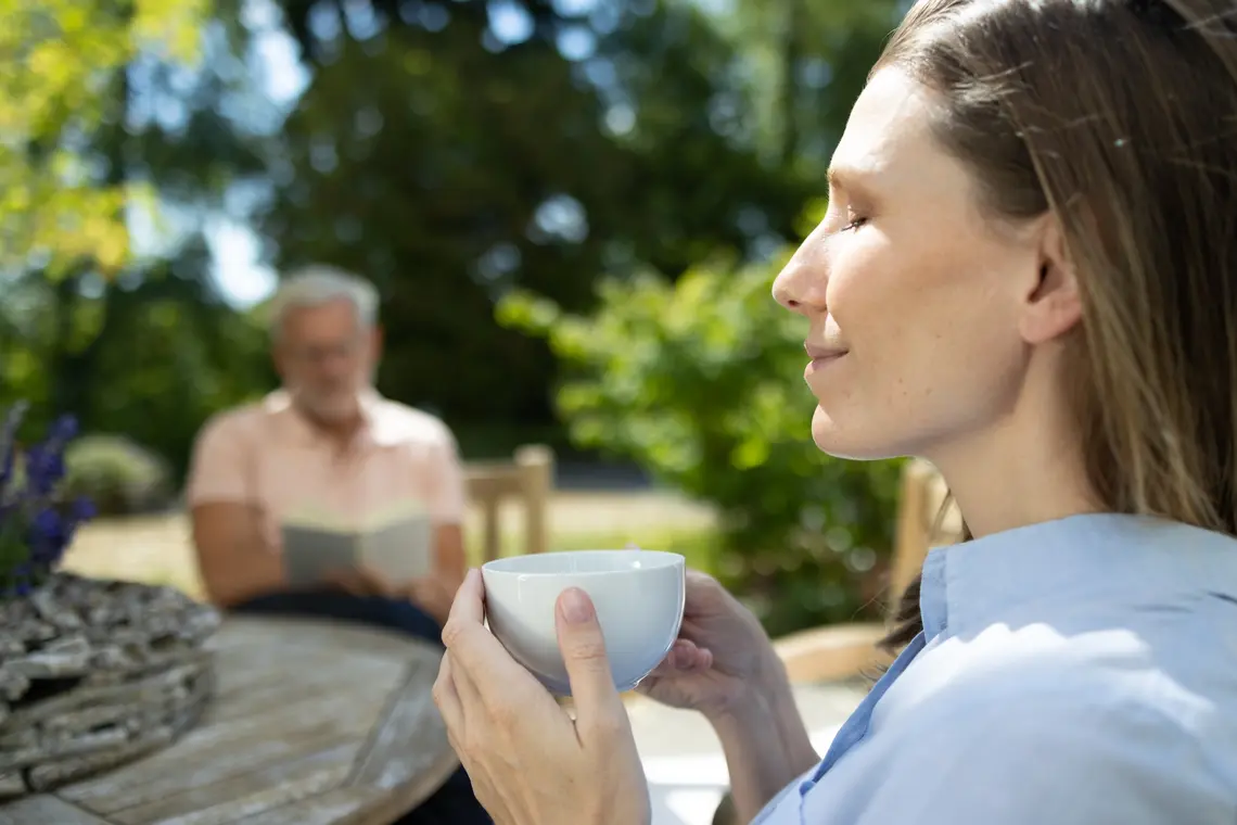 Eine Frau hält eine Tasse in beiden Händen. Im Hintergrund liest ein Mann ein Buch. Sie sitzen draußen an einem Tisch.