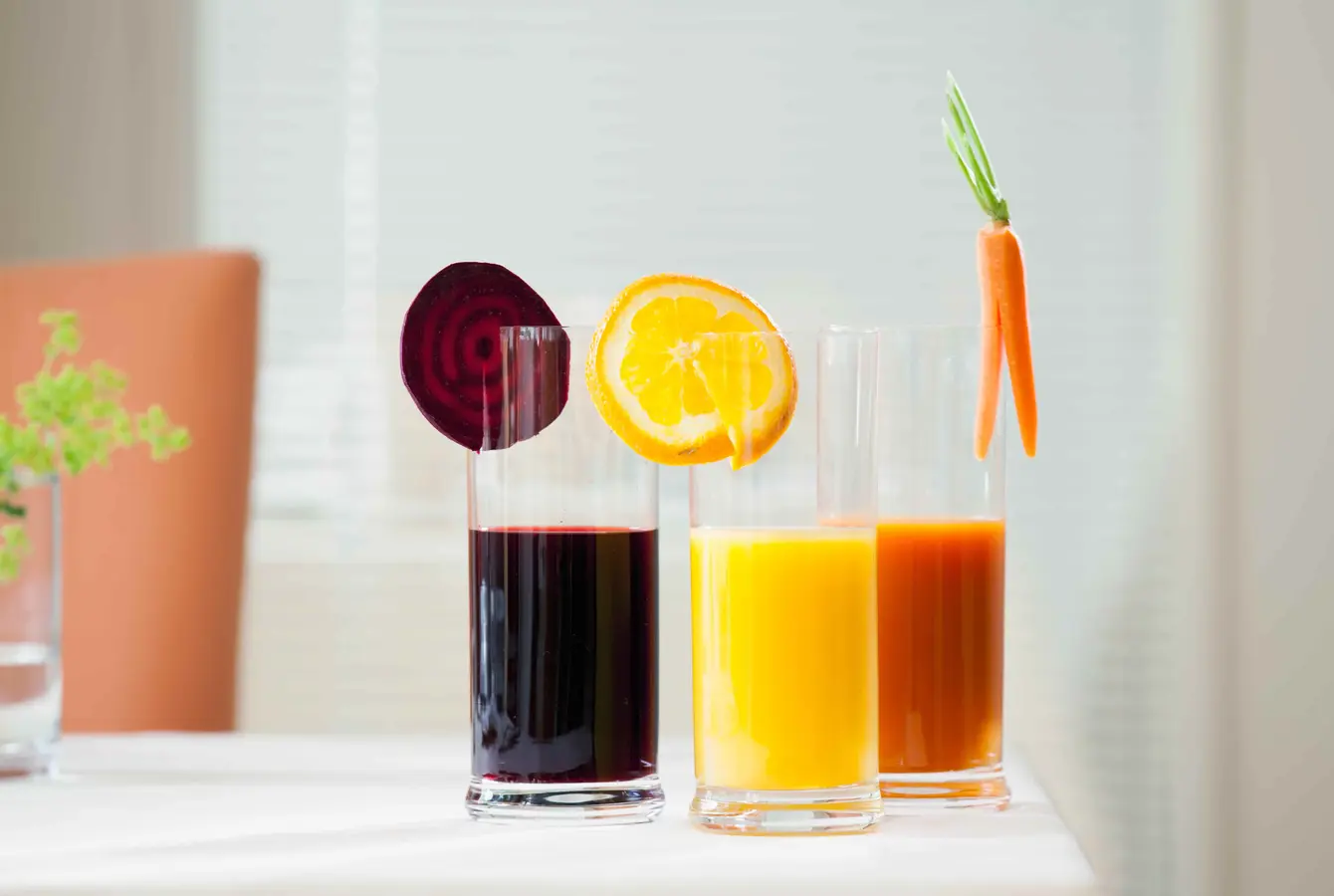 Drei Gläser Saft stehen nebeneinander auf einem weißen Tisch. Rote Bete, Orange und Karotte hängt jeweils in einem Saftglas