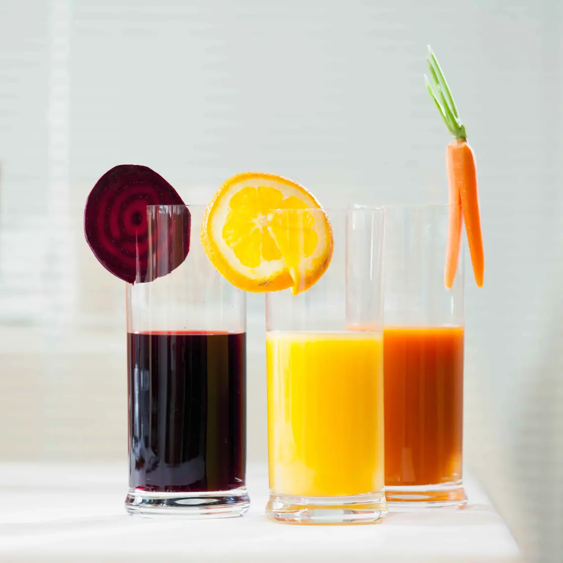 Drei Gläser Saft stehen nebeneinander auf einem weißen Tisch. Rote Bete, Orange und Karotte hängt jeweils in einem Saftglas