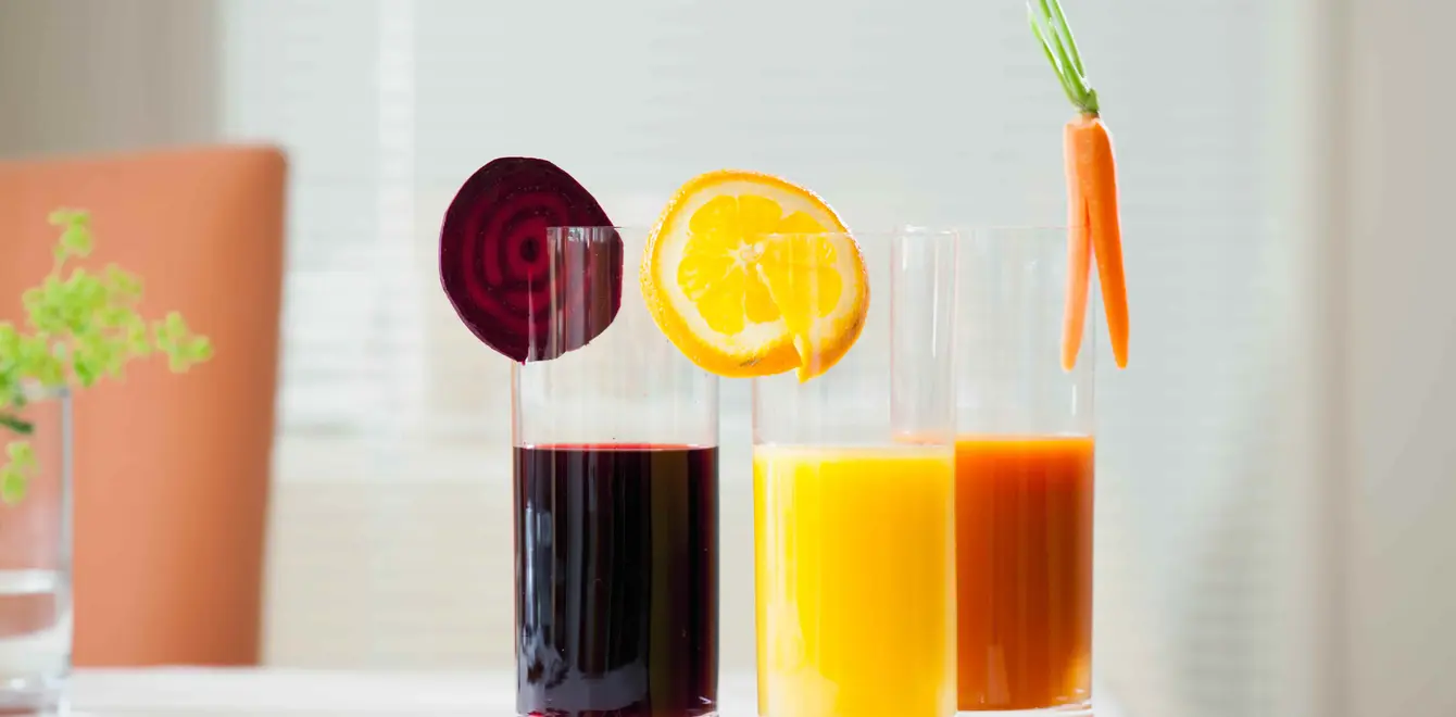 Drei Gläser Saft stehen nebeneinander auf einem weißen Tisch. Rote Bete, Orange und Karotte hängt jeweils in einem Saftglas