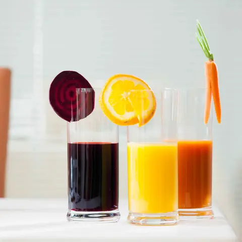 Drei Gläser Saft stehen nebeneinander auf einem weißen Tisch. Rote Bete, Orange und Karotte hängt jeweils in einem Saftglas