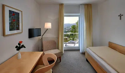 Helles Zimmer mit Einzelbett, Schreibtisch mit Stuhl, Sessel und Fernseher. Blick auf Balkontür.
