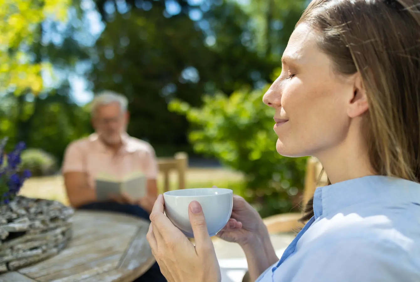 Eine Frau sitzt im Garten der Dr. von Weckbecker Klinik, hält mit beiden Händen eine Tasse vor sich und hat entspannt die Augen geschlossen. Im Hintergrund sitzt ein Mann auf der Wiese und liest ein Buch.