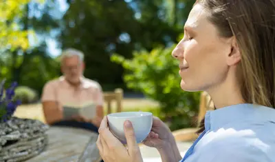 Eine Frau sitzt im Garten der Dr. von Weckbecker Klinik, hält mit beiden Händen eine Tasse vor sich und hat entspannt die Augen geschlossen. Im Hintergrund sitzt ein Mann auf der Wiese und liest ein Buch.