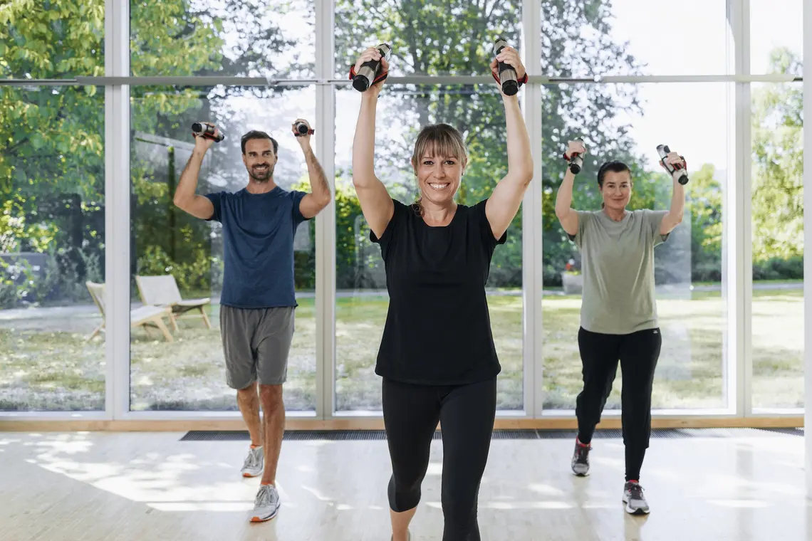 Zwei Frauen und ein Mann beim Sport. Sie tragen Fitness-Gewicht in den Händen über den Schultern