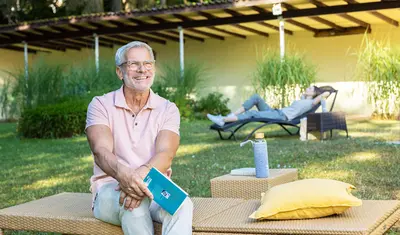 Ein Mann sitzt auf einer Gartenliege auf der Liegewiese der Dr. von Weckbecker klinik. Er hält ein Buch in der Hand und schaut Richtung Sonne. Im Hintergrund liegt eine Frau auf einer liege auf der selben Wiese.