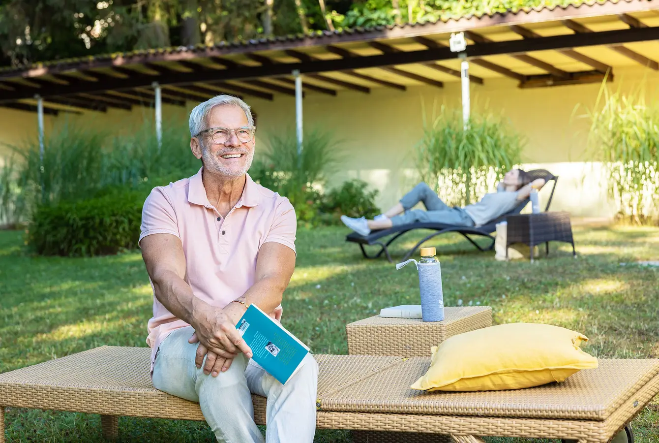 Ein Mann sitzt auf einer Gartenliege auf der Liegewiese der Dr. von Weckbecker klinik. Er hält ein Buch in der Hand und schaut Richtung Sonne. Im Hintergrund liegt eine Frau auf einer liege auf der selben Wiese.
