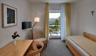 Helles Zimmer mit Einzelbett, Schreibtisch mit Stuhl, Sessel und Fernseher. Blick auf Balkontür.
