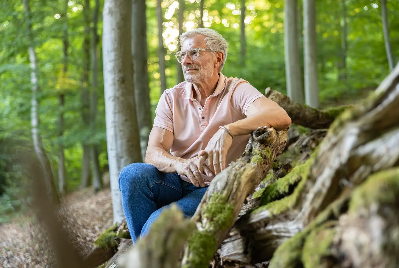 Das Bild zeigt einen Mann welcher, angelehnt an einen liegenden Baumstamm, im Wald sitzt und in die Ferne schaut.