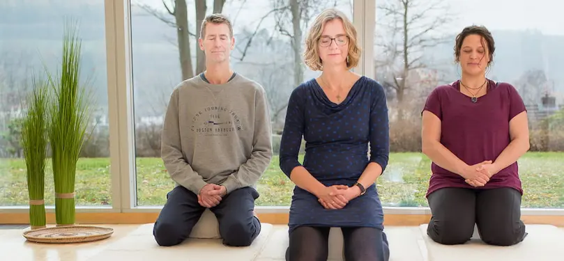 Ein Mann und zwei Frauen Meditieren auf den Knien vor einer großen Fensterfront