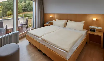 Doppelbett mit weißen Bezüge und Gestell in Holzoptik. Blick zur Balkontür mit Sitzgelegheiten.