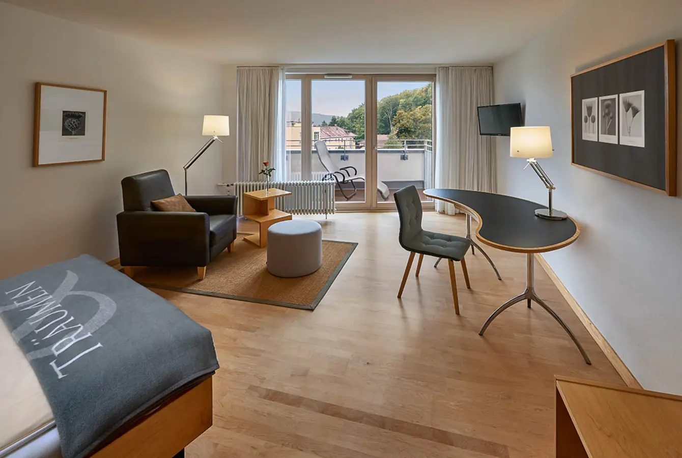 Modernes geräumiges Einzelzimmer mit Bett, Ledersessel, gebogenem Schreibtisch und Schreibtischstuhl. Fernsehen und großer Balkontür und Fensterfront.