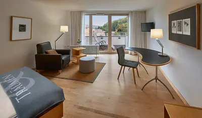 Modernes geräumiges Einzelzimmer mit Bett, Ledersessel, gebogenem Schreibtisch und Schreibtischstuhl. Fernsehen und großer Balkontür und Fensterfront.