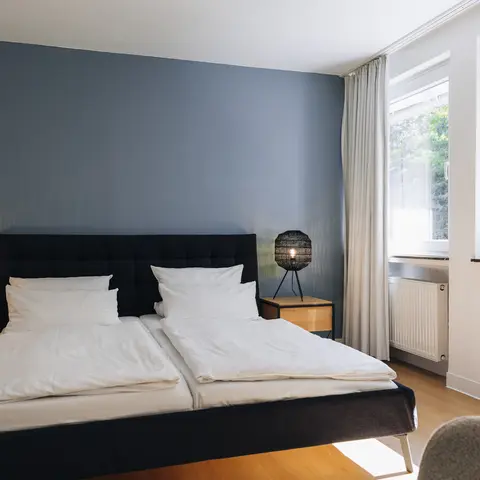 Schwarzes Doppelbett mit weißen Bezügen und zwei Nachttischlampen vor einer blauen Wand. Blick aus dem Fenster ins Grüne.
