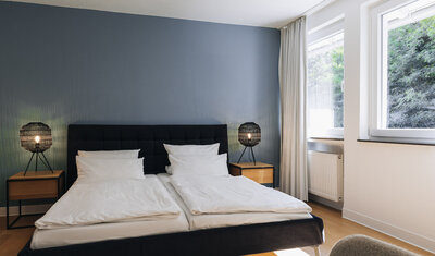 Schwarzes Doppelbett mit weißen Bezügen und zwei Nachttischlampen vor einer blauen Wand. Blick aus dem Fenster ins Grüne.