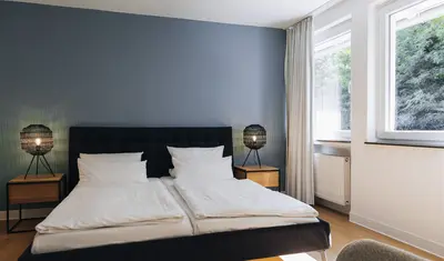 Schwarzes Doppelbett mit weißen Bezügen und zwei Nachttischlampen vor einer blauen Wand. Blick aus dem Fenster ins Grüne.