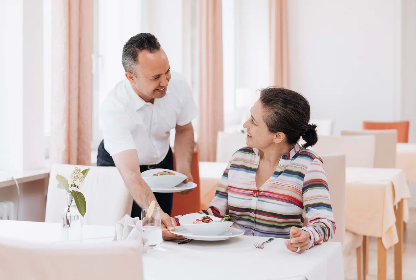 Das Bild zeigt eine Szene im Speisesaal der Dr. von Weckbecker Klinik. Eine Frau sitzt an einem Tisch und bekommt von einem Kellner einen Teller mit Essen auf den Tisch gestellt.
