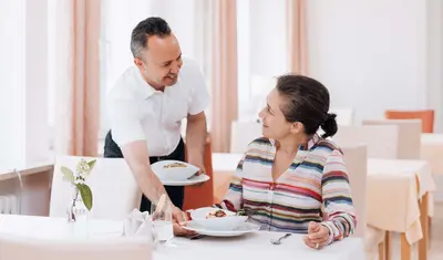 Das Bild zeigt eine Szene im Speisesaal der Dr. von Weckbecker Klinik. Eine Frau sitzt an einem Tisch und bekommt von einem Kellner einen Teller mit Essen auf den Tisch gestellt.