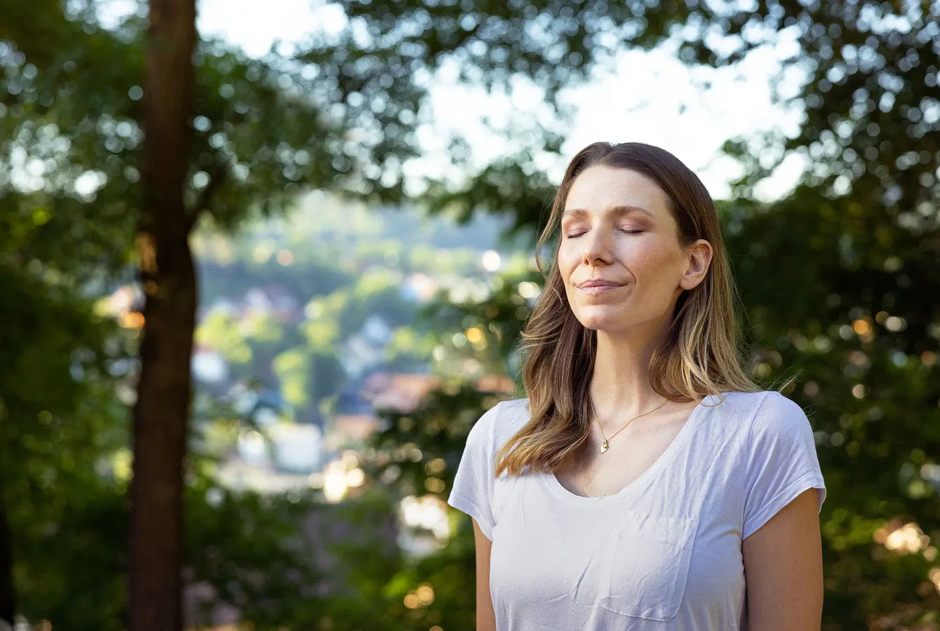 Auf dem Bild sieht man eine Frau, welche mit verschlossenen Augen, tief luftholend inmitten der Natur steht. Im Hintergrund sieht man eine Baumreihe.
