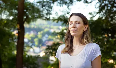 Auf dem Bild sieht man eine Frau, welche mit verschlossenen Augen, tief luftholend inmitten der Natur steht. Im Hintergrund sieht man eine Baumreihe.