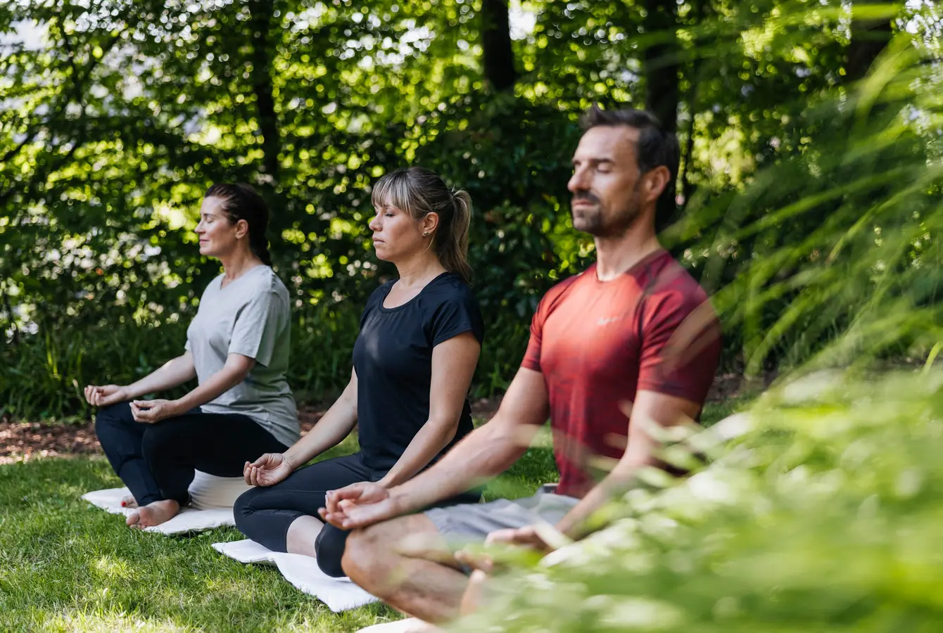 Zwei Frauen und ein Mann meditieren nebeneinander auf einer Wiese im Schatten von Bäumen