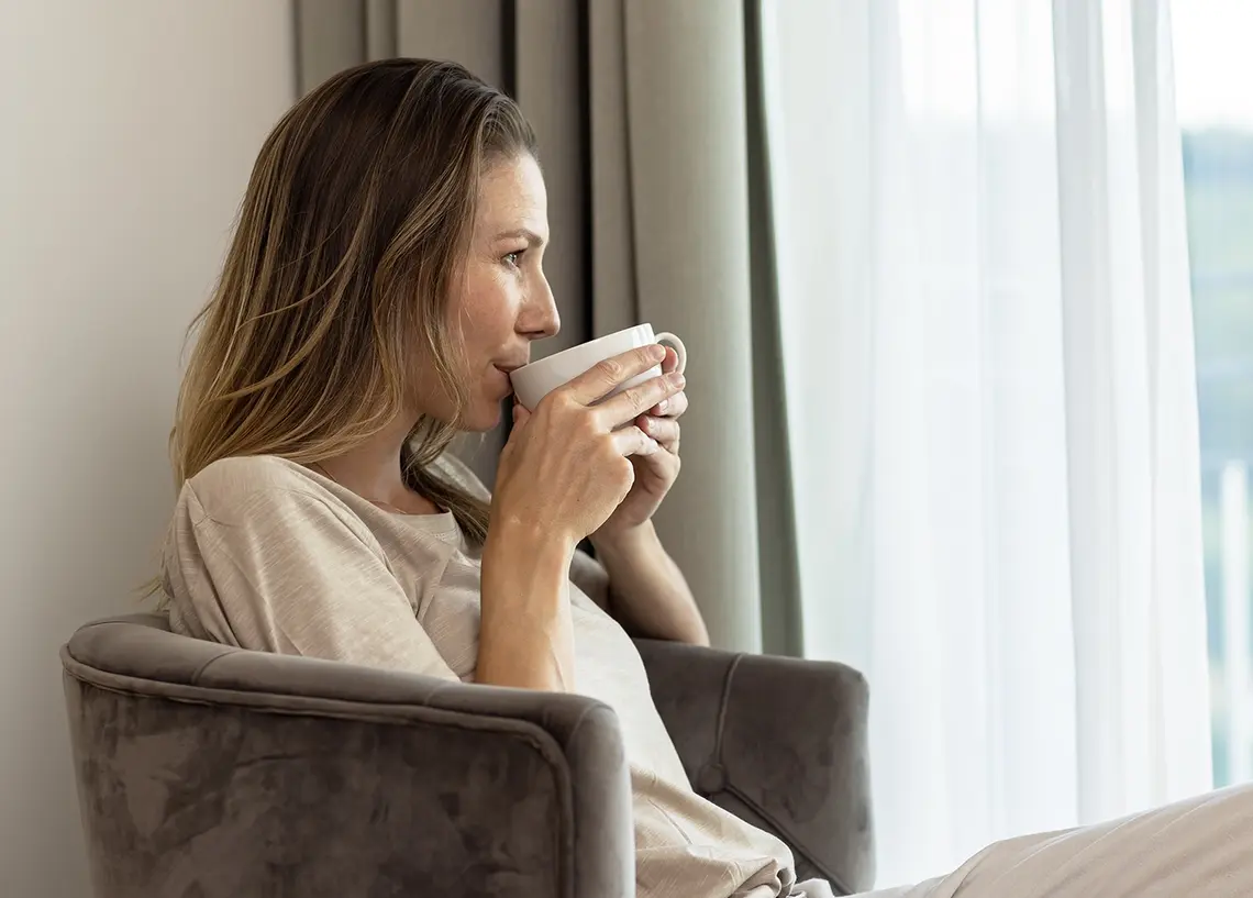 Eine Frau trinkt aus eine Tasse, sitzt in einem Sessel und schaut aus dem Fenster