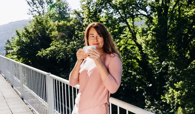 Eine Frau steht auf einem Balkon der Dr. von Weckbecker Klinik, lehnt sich ans Geländer an und hält eine Tasse vor dem Gesicht. Im Hintergrund sieht man den Rest des Balkons und die Bäume am Grundstücksrand.