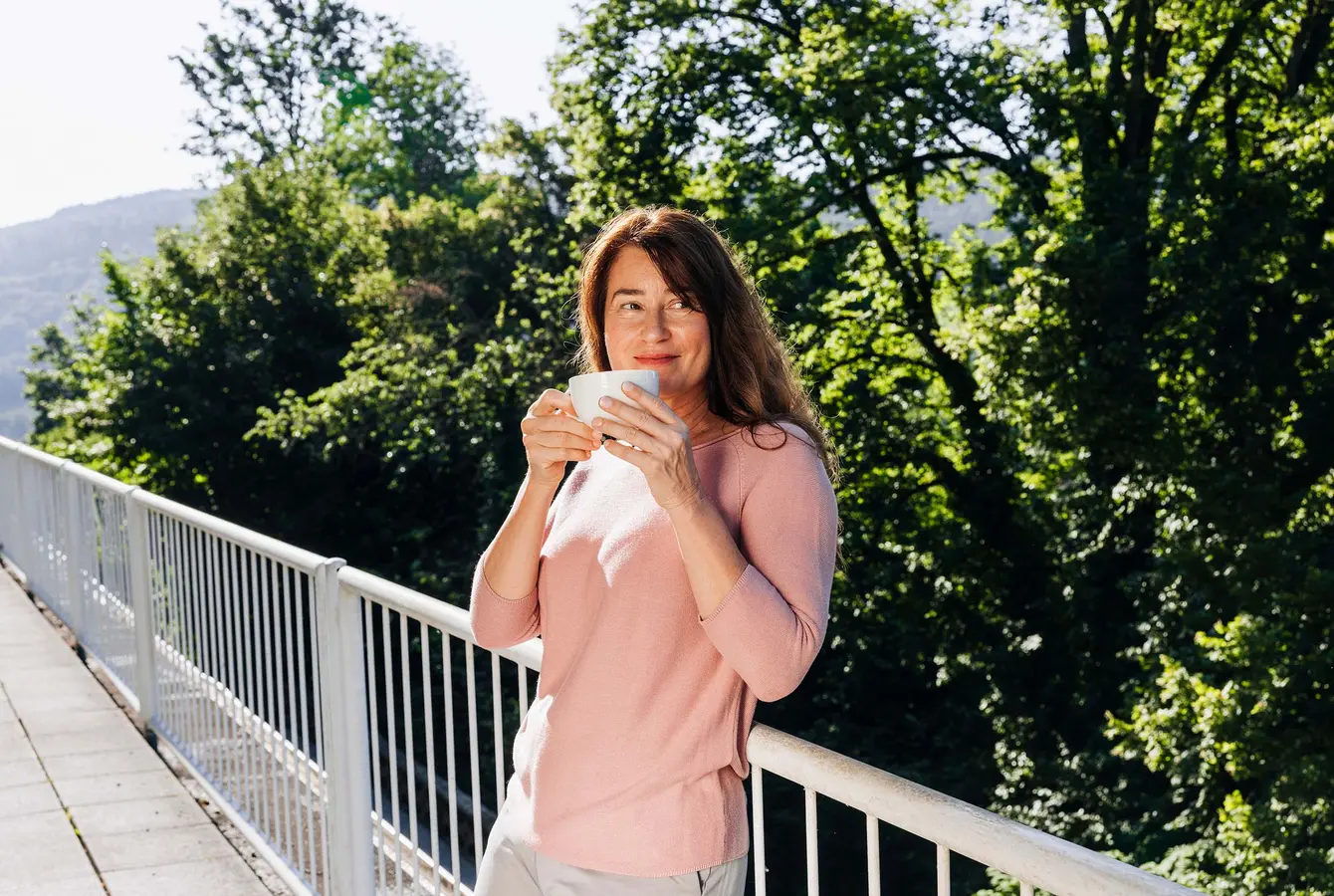 Eine Frau steht auf einem Balkon der Dr. von Weckbecker Klinik, lehnt sich ans Geländer an und hält eine Tasse vor dem Gesicht. Im Hintergrund sieht man den Rest des Balkons und die Bäume am Grundstücksrand.